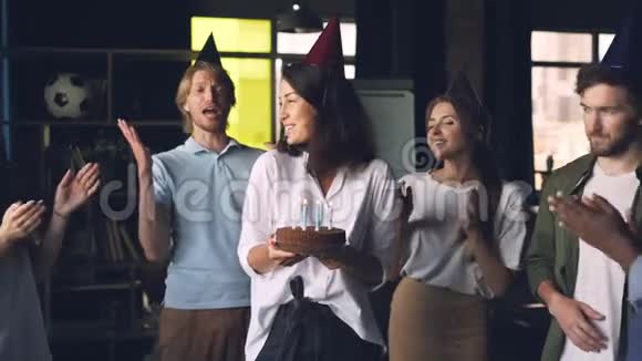 年轻的女士拿着生日蛋糕吹着蜡烛站在办公室里同事们鼓掌视频的预览图