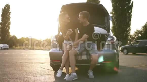 快乐的沙发坐在敞篷的汽车后备箱里戴着死锁和太阳镜的漂亮女孩在智能手机上滚动视频的预览图