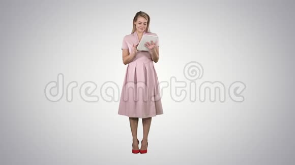 漂亮的年轻女人穿着粉红色的连衣裙拿着平板电脑在渐变的背景下看着它视频的预览图