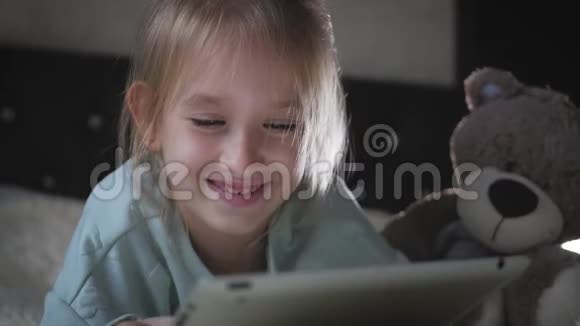 可爱的小女孩坐在床上和泰迪熊一起在平板电脑上玩游戏视频的预览图