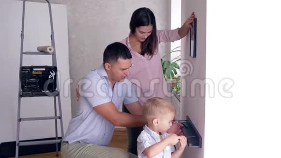 爸爸在公寓装修期间教儿子用螺丝刀把架子栓到墙上妈妈在附近挂画视频的预览图