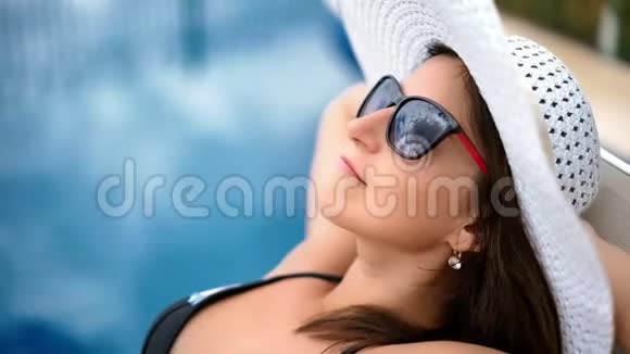 戴着太阳镜和帽子享受日光浴的漂亮女人躺在游泳池附近的躺椅上视频的预览图