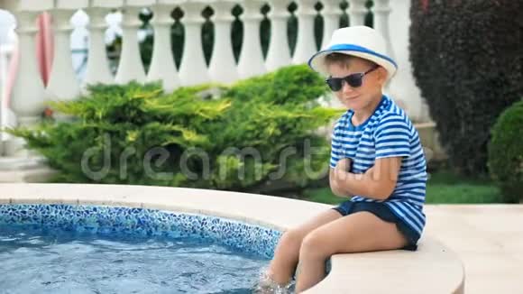 动作缓慢的小男孩戴着太阳镜和帽子坐在游泳池的边缘在水中玩腿视频的预览图