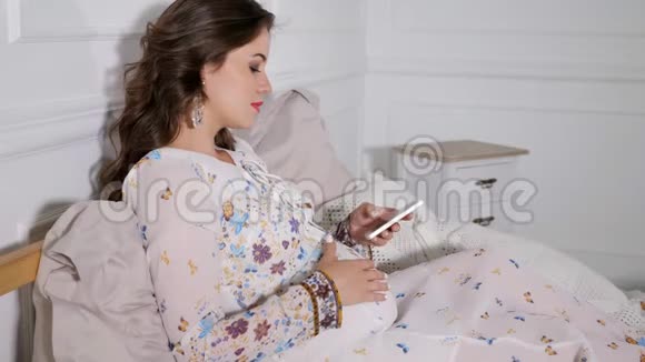 在家里坐在床上用智能手机的孕妇微笑视频的预览图