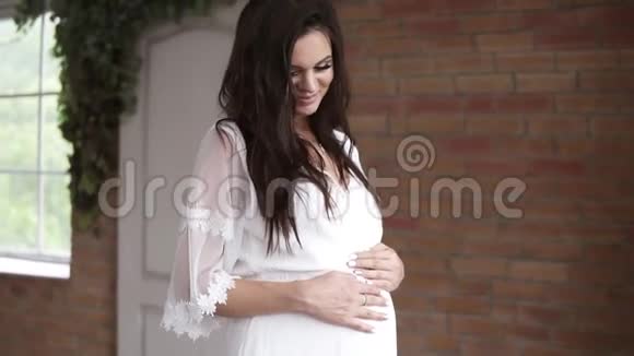 穿着白色连衣裙的孕妇在棕色砖墙背景上摸着她的肚子微笑美丽的黑发女人视频的预览图