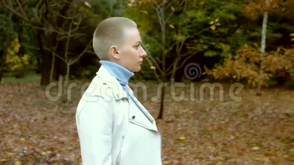 时尚的金发女人穿着白色的皮夹克留着很短的头发走在秋天的公园里看着摄像机视频的预览图