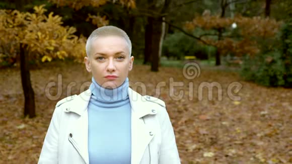 时尚的金发女人穿着白色的皮夹克留着很短的头发走在秋天的公园里看着摄像机视频的预览图