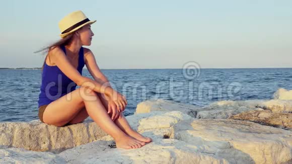 穿着蓝色T恤和巴拿马短裤的女孩坐在海边的白色石头上视频的预览图