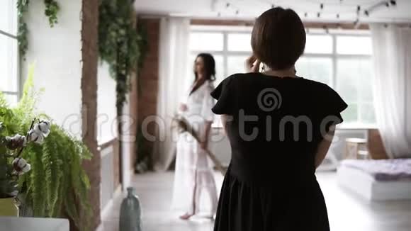 从后面的录像女摄影师穿着黑色连衣裙为美丽的孕妇拍照灯光装饰视频的预览图