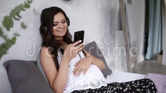 年轻孕妇坐在沙发上布鲁内特微笑的女士长发抚摸着她的腹部看着她视频的预览图
