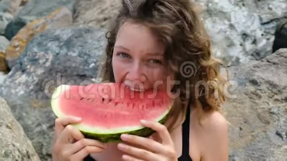 一个年轻漂亮的卷发女人咬掉大海上的西瓜的肖像视频的预览图