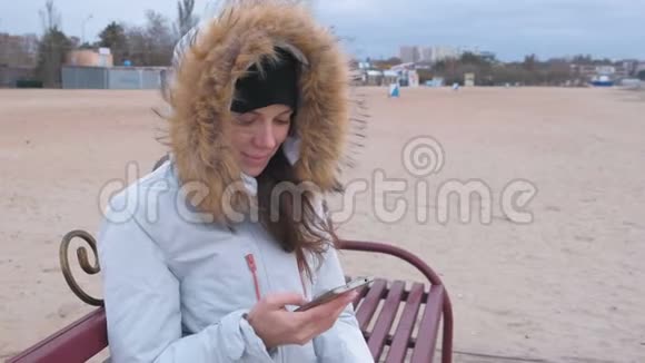 穿着白色羽绒服的女人坐在海滩上的长凳上看着手机看着令人惊叹的视频视频的预览图
