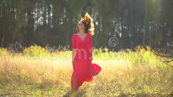 美丽浪漫微笑的年轻女子穿着红色长裙在夕阳下在森林里跳舞美丽快乐的女模特女孩视频的预览图