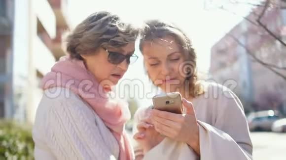 母亲和成年女儿在阳光明媚的风中站在春街的中央用智能手机看照片视频的预览图