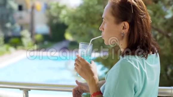 一位年轻女子站在阳台上戴上太阳镜用吸管喝一杯鸡尾酒4k视频的预览图