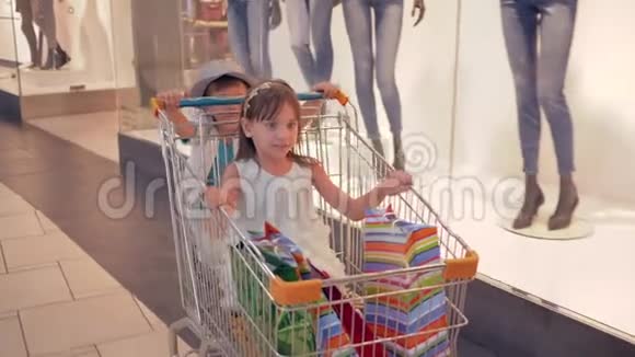 儿童购物日情绪活跃的孩子在商场的顾客手推车里玩得开心从商店橱窗里走过视频的预览图
