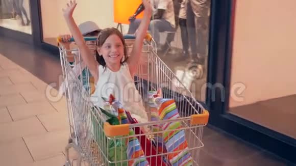 折扣季节欢笑的孩子们在商场的购物车里玩得很开心从精品店的橱窗里走过视频的预览图