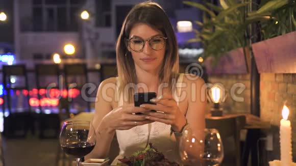 优雅的女人用手机坐在晚上的餐厅的餐桌上漂亮的女人看着智能手机视频的预览图
