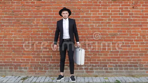 一个年轻的白种人穿着黑色西装打开一个金属手提箱在砖墙背景下带着钱动作缓慢视频的预览图
