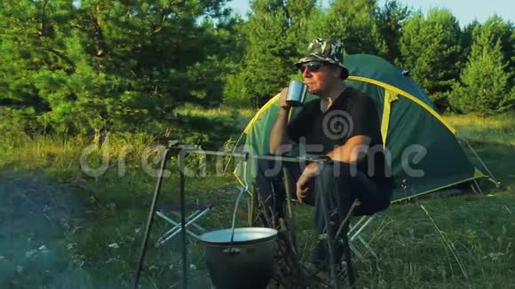 戴墨镜的人坐在帐篷里喝茶看着挂在炉火上的保龄球视频的预览图