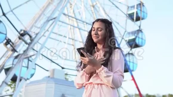 一个留着深色长发的女孩在一个游乐园里一个站在大摩天轮旁的智能手机心情很好视频的预览图