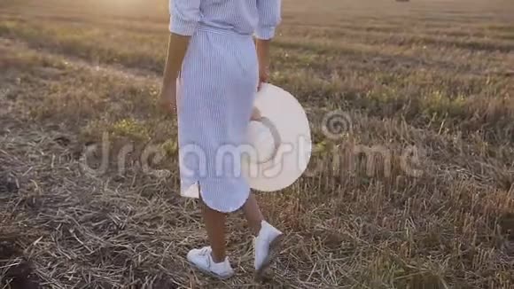 夏天一个身材苗条的女孩穿着一条蓝色的长裙子手里拿着一顶大帽子走在金色的田野里视频的预览图
