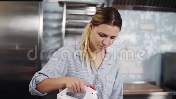 一个穿着蓝色衬衫和黑色围裙的糖果者用搅拌器将蛋清在钢碗中敲打成奶油状视频的预览图