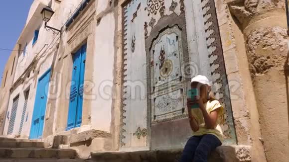 年轻女孩使用电话坐在老城区传统阿拉伯门附近的台阶上视频的预览图