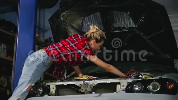 可爱的金发女孩修理汽车并在服务站的汽车罩下用帮助扳手固定发动机的细节视频的预览图