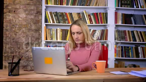 集中的高加索女孩的肖像带着穿心工作与笔记本电脑在书架背景视频的预览图