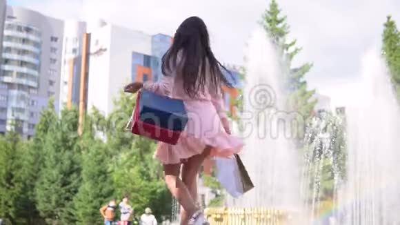 一个女孩在公园的喷泉附近购物后手里拿着袋子慢动作高清视频的预览图