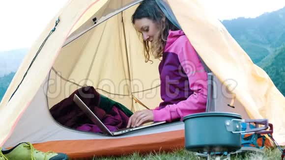 快乐的旅游女孩坐在帐篷里用笔记本电脑自由职业者在旅行中在大自然上打印视频的预览图