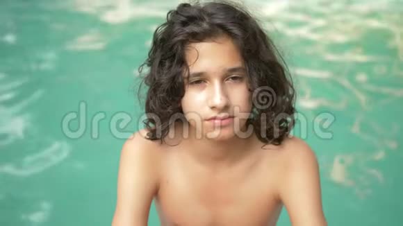 一个微笑的少年男孩一头卷曲的黑发坐在游泳池旁躺在蓝色的水背景上视频的预览图