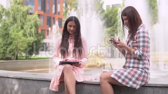 两个女学生坐在喷泉附近的公园里用智能手机和平板电脑慢动作视频的预览图
