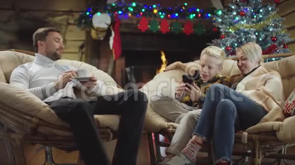 父亲喝咖啡而他的妻子和儿子在圣诞夜看智能手机视频视频的预览图