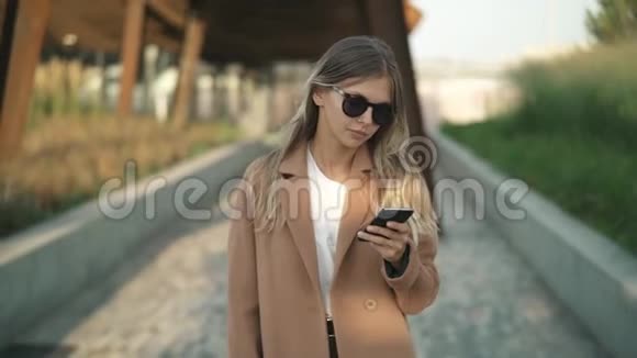 年轻的金发女郎走在公园里看着智能手机然后微笑着视频的预览图