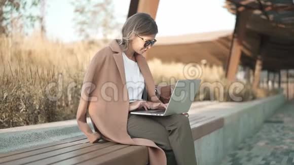 一位身着米色外套带着笔记本电脑在公园工作的冷静金发女人视频的预览图