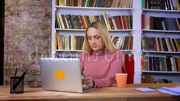 集中的白种人年轻女孩的肖像穿着穿孔专心工作笔记本电脑在书架背景视频的预览图