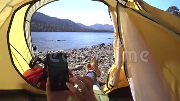 女旅行者躺在湖边的一个黄色帐篷里手里拿着智能手机然后是视频的预览图
