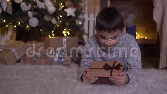 一个小男孩打开一个带礼物的盒子高兴地躺在圣诞树附近的地板上4K视频的预览图