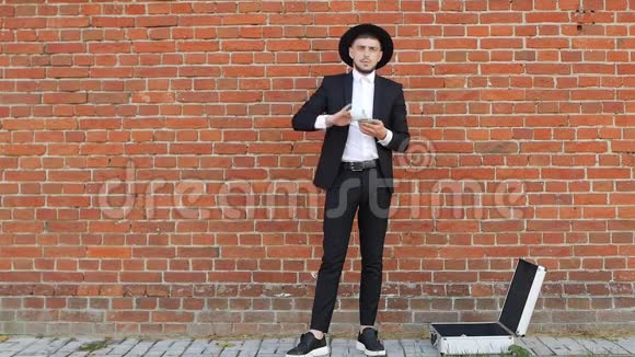 一个穿着黑色西装戴着帽子的年轻人在砖墙背景上算钱动作慢风格好视频的预览图