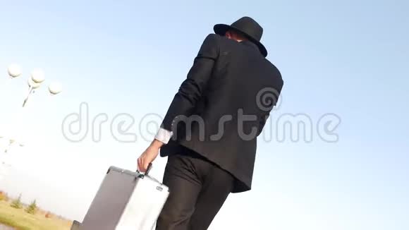 一个穿着黑色时髦西装戴着帽子的年轻人带着手提箱用手机说话动作缓慢视频的预览图
