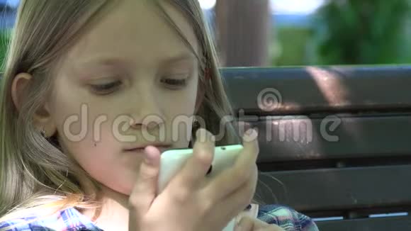 儿童玩智能手机在公园的游乐场放桌女孩坐在长凳上视频的预览图