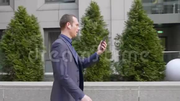 一位年轻的商人用无线耳机走在他的耳朵里用智能手机视频通话视频的预览图