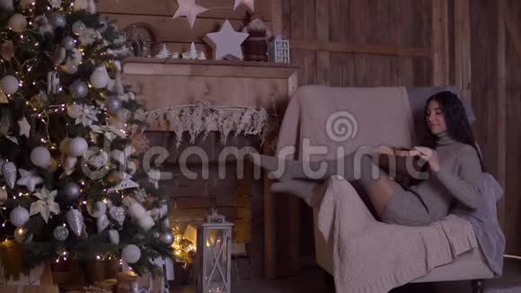 一个留着长发的女孩打开一个装有礼物的盒子并在圣诞节的时候坐在圣诞树附近的椅子上高兴视频的预览图