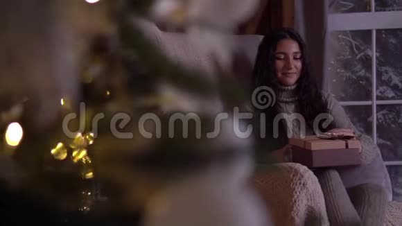 女孩拿着礼物打开盒子坐在圣诞树附近的椅子上高兴高清高清视频的预览图