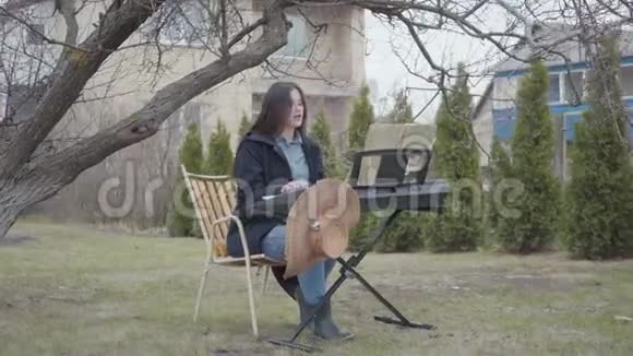 聪明漂亮的深褐色头发的女孩在树下坐着热情地玩合成器唱歌视频的预览图