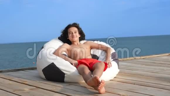 英俊的少年一头卷发戴着太阳镜坐在海边的一个木制露台上的包椅里概念视频的预览图