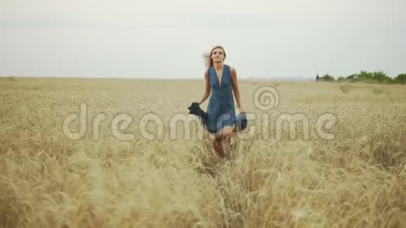 英俊的年轻女子穿着一件蓝色的长裙穿过金色的麦田看着镜头自由概念视频的预览图