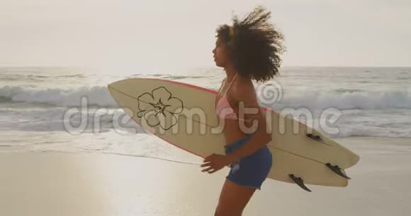 非洲裔美国女性冲浪者在海滩上用冲浪板跑步的侧面景色视频的预览图